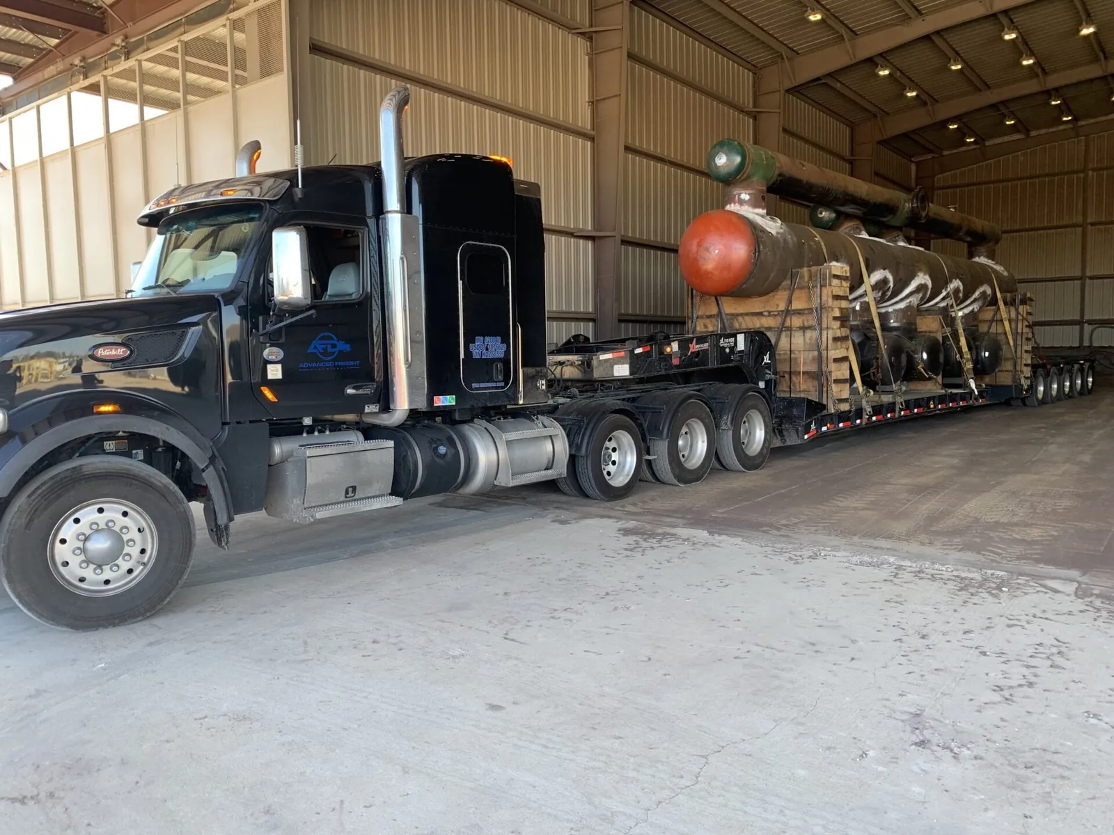 Black semi truck with an oversize load parked inside a large shop building