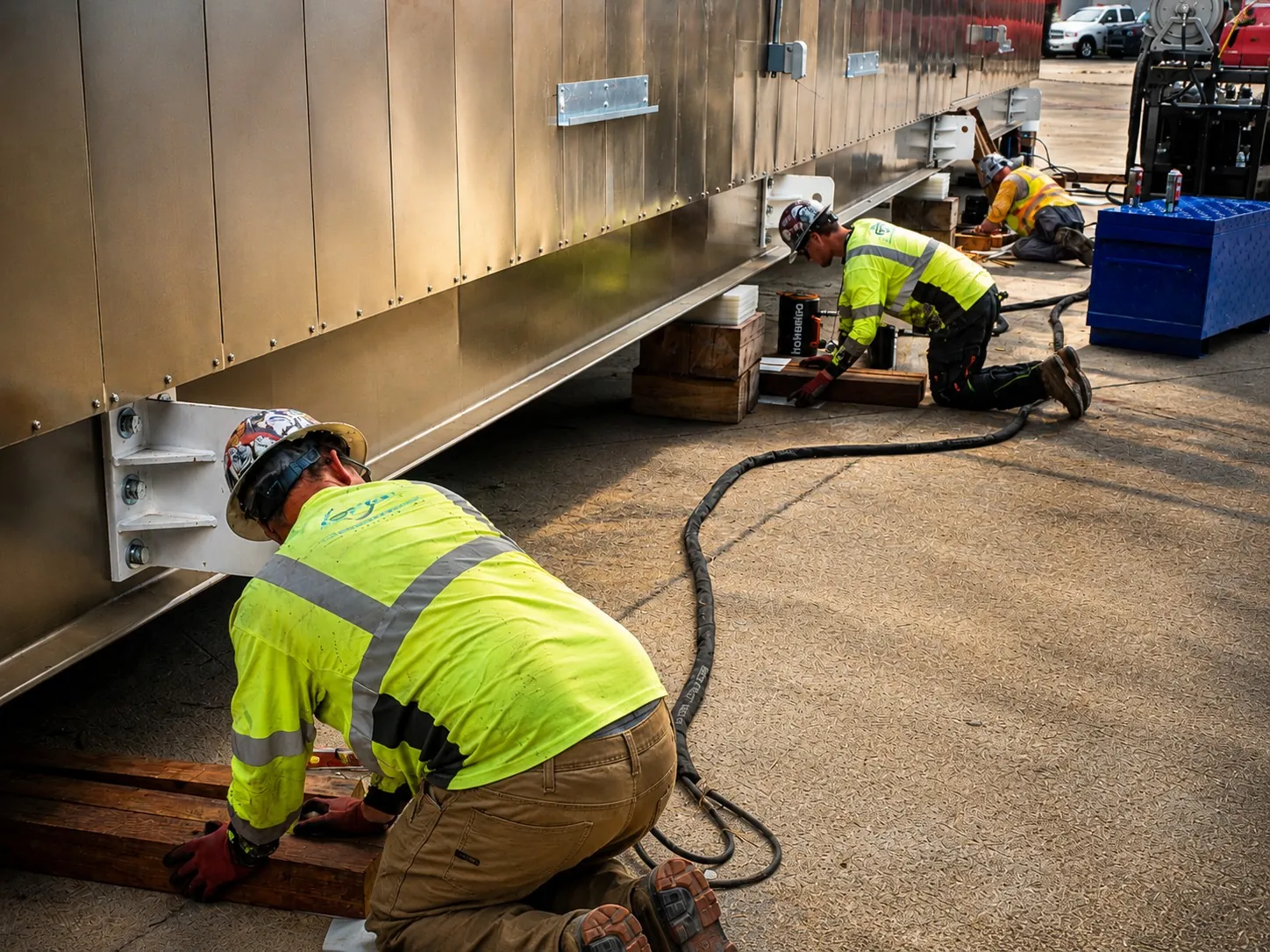 Crew in hi-vis vests setting a metal-clad prefab power distribution building on wood cribbing